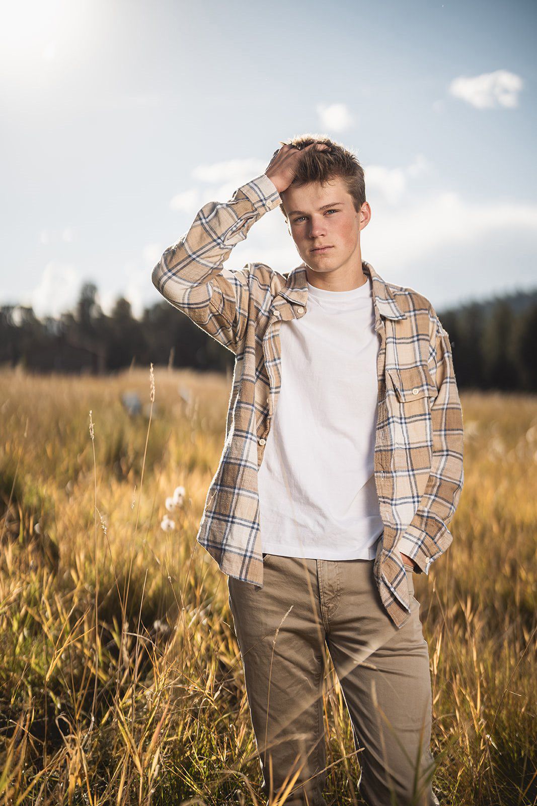 Teen boy in plaid shirt and white tee standing in golden field with hand in hair.