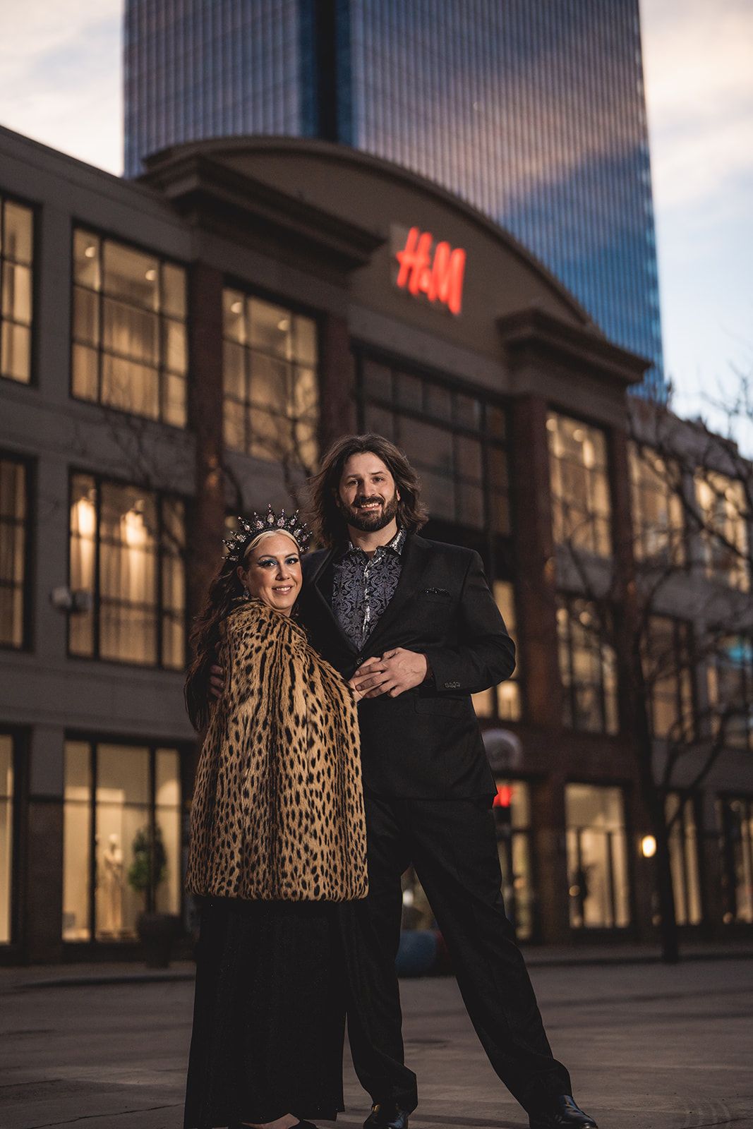 Couple posing in front of H&M store at dusk, woman in leopard coat with crown, man in black suit.