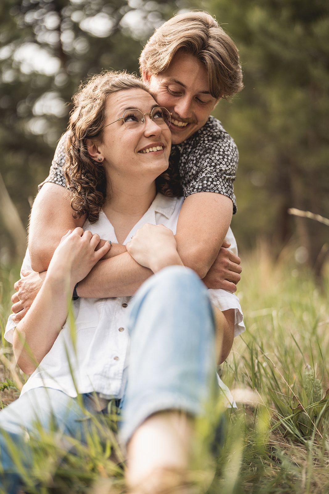 A couple sitting in grass, man embracing woman from behind as she smiles up at him in a sunny outdoor setting.