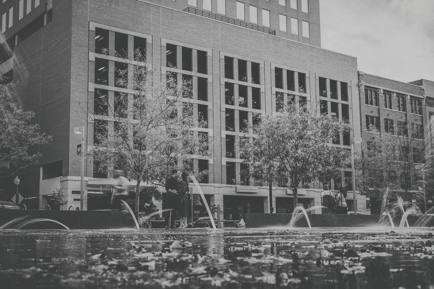 Black and white photo of a modern brick building with large windows, trees, and a fountain in the foreground.