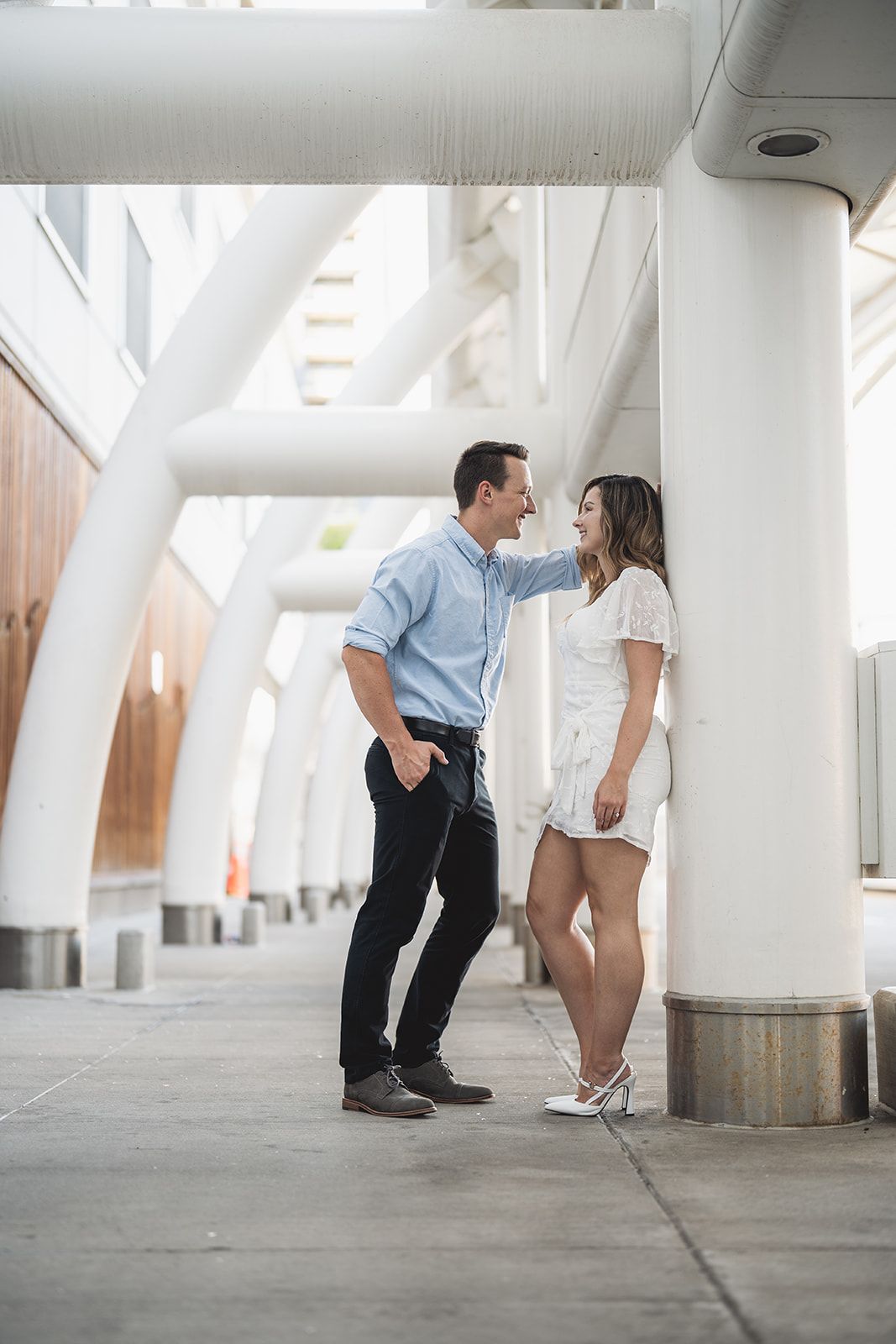 A couple shares a romantic moment leaning against white columns in a modern urban setting, man in blue shirt, woman in white dress.
