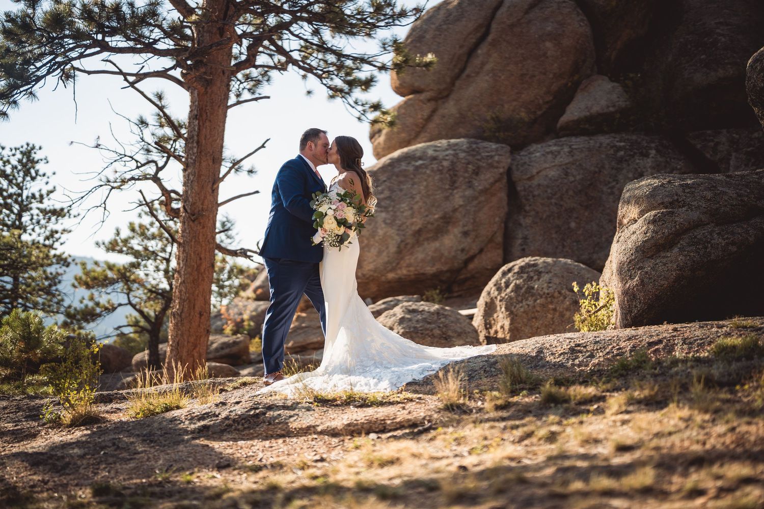 Couple posing on rocky mountain outcrop surrounded by pine trees at sunset.