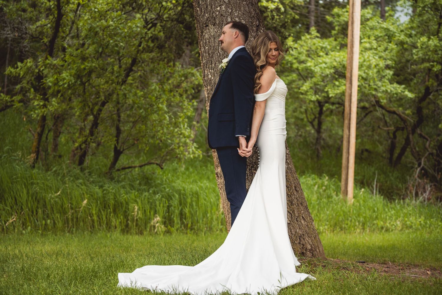 Newlyweds in white dress and navy suit pose romantically against forest backdrop.