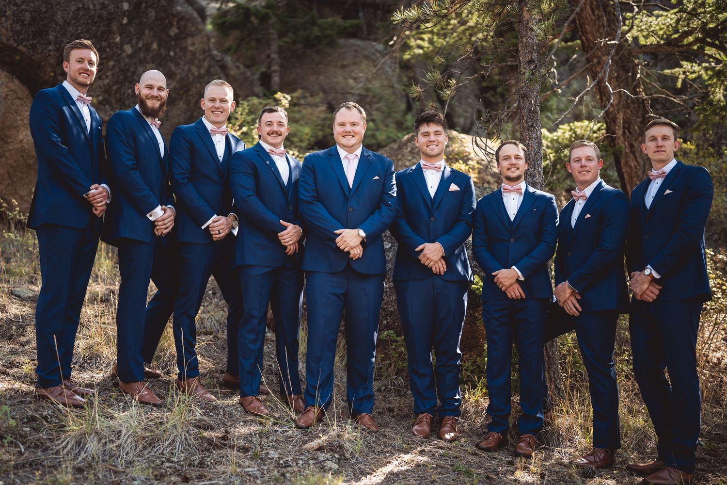 Wedding party members in navy suits line up along a natural mountain trail.