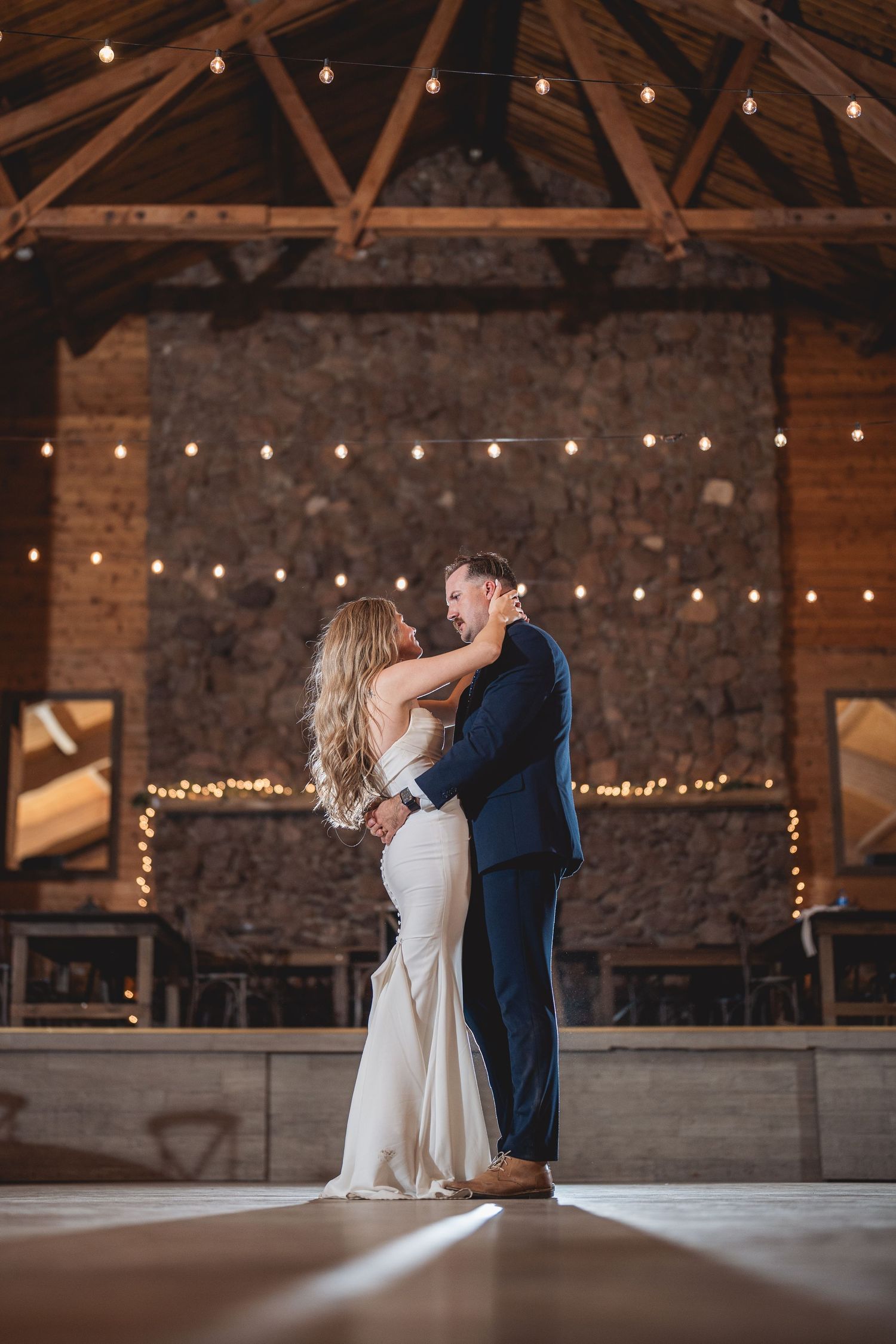 A couple shares an intimate dance moment in a rustic barn with string lights and exposed brick walls.