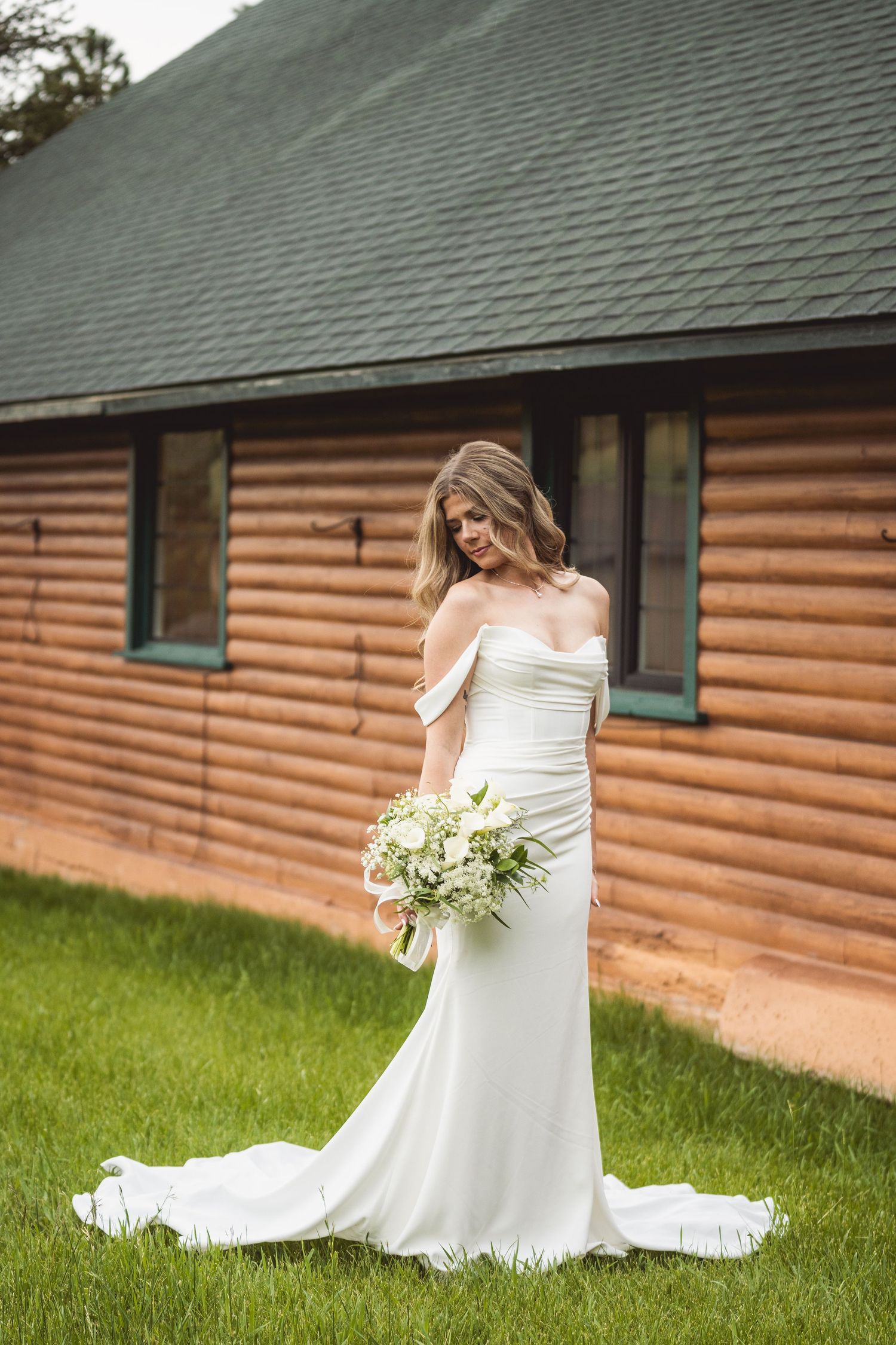 A white wedding dress train flows across green grass near a rustic log cabin.