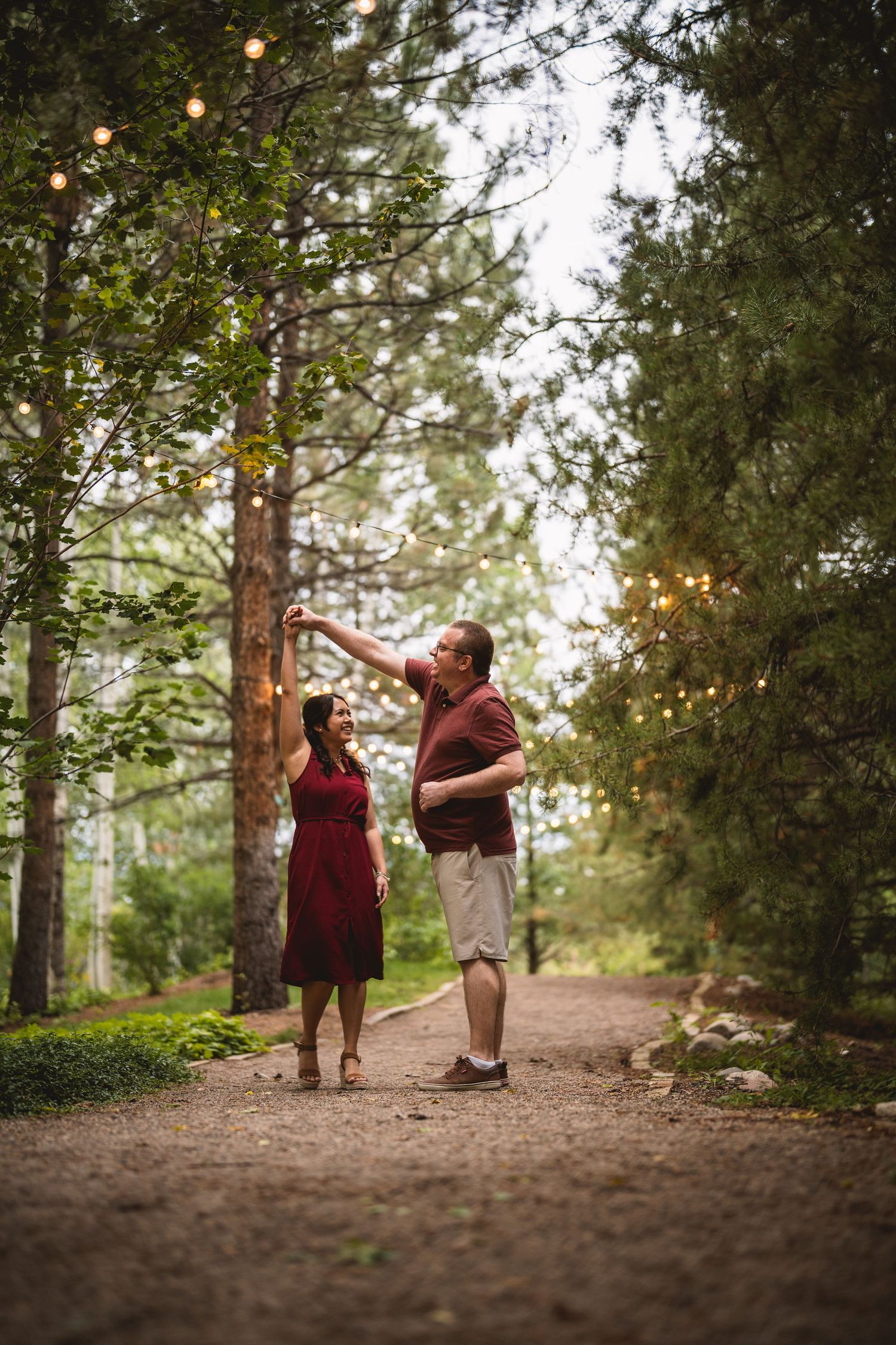 A couple walks along a wooded path with string lights hanging overhead at dusk.