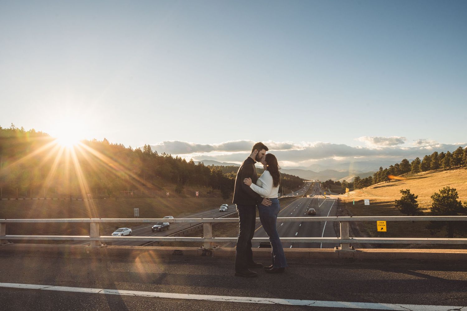 Couple embracing at scenic mountain viewpoint while sun sets behind clouds.