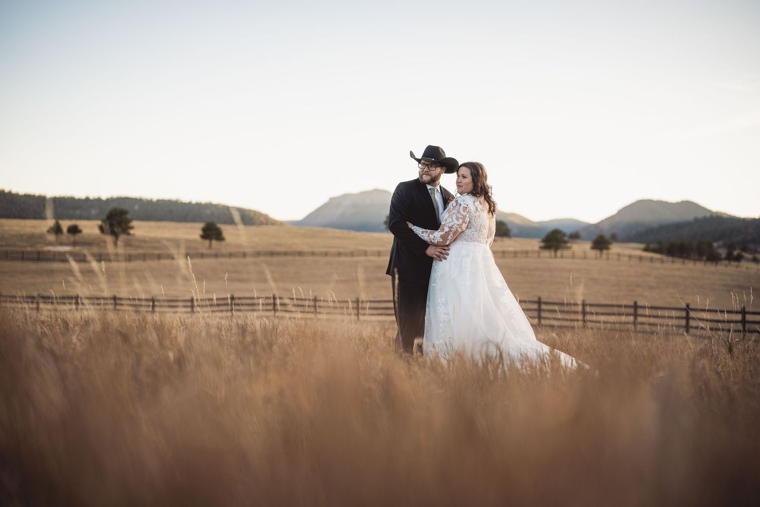 Wedding couple embracing in golden field with mountain backdrop and rustic fencing.
