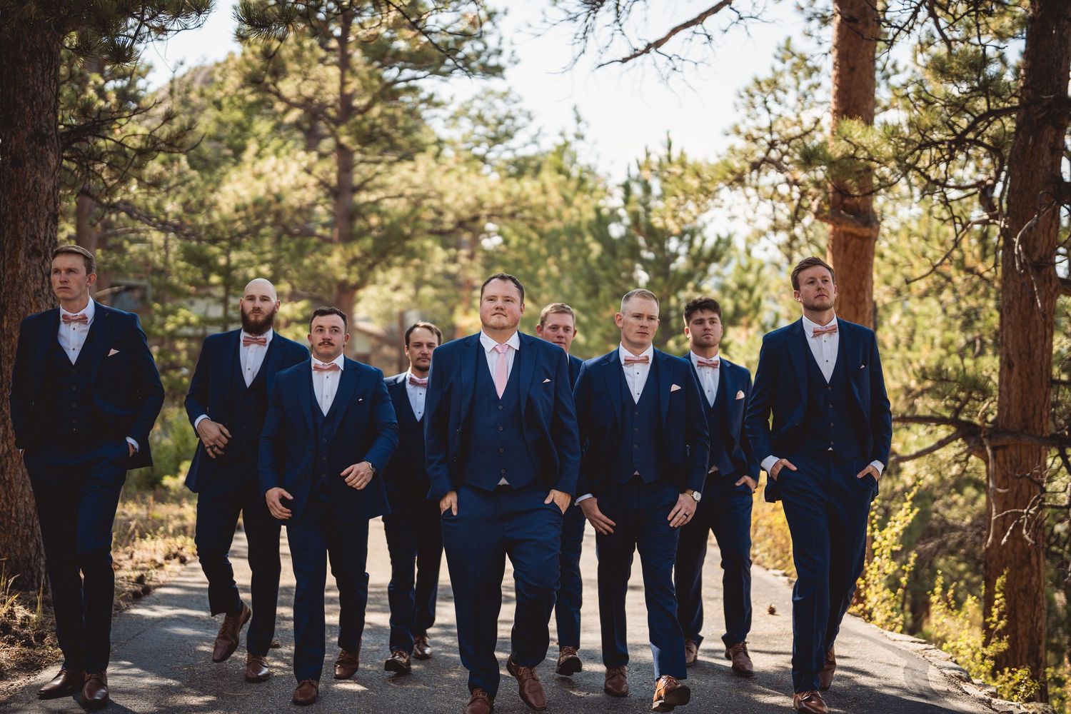 A wedding party in navy suits poses together outdoors in a wooded mountain setting.