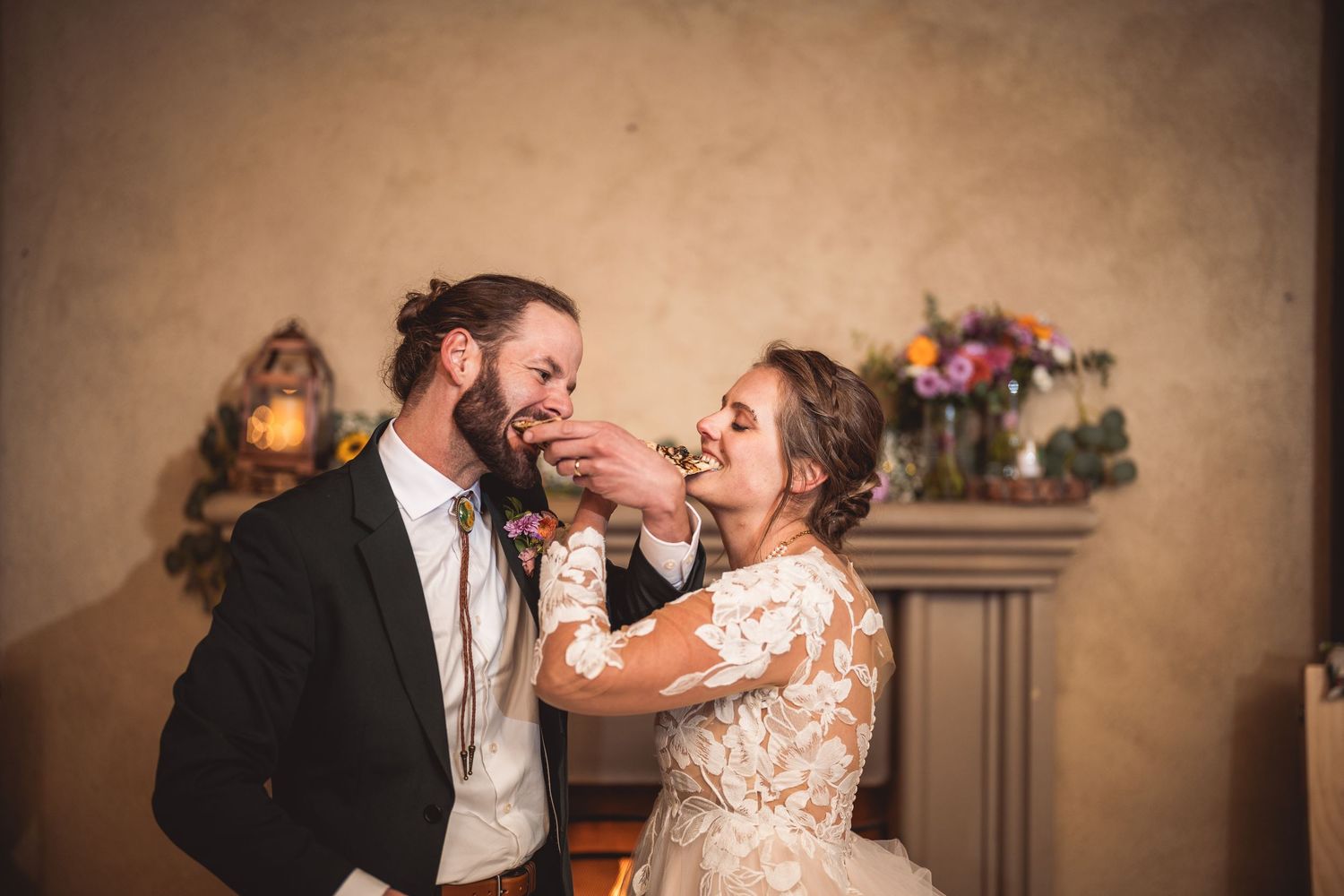 Couple shares a playful cake-feeding moment in front of a decorated fireplace.