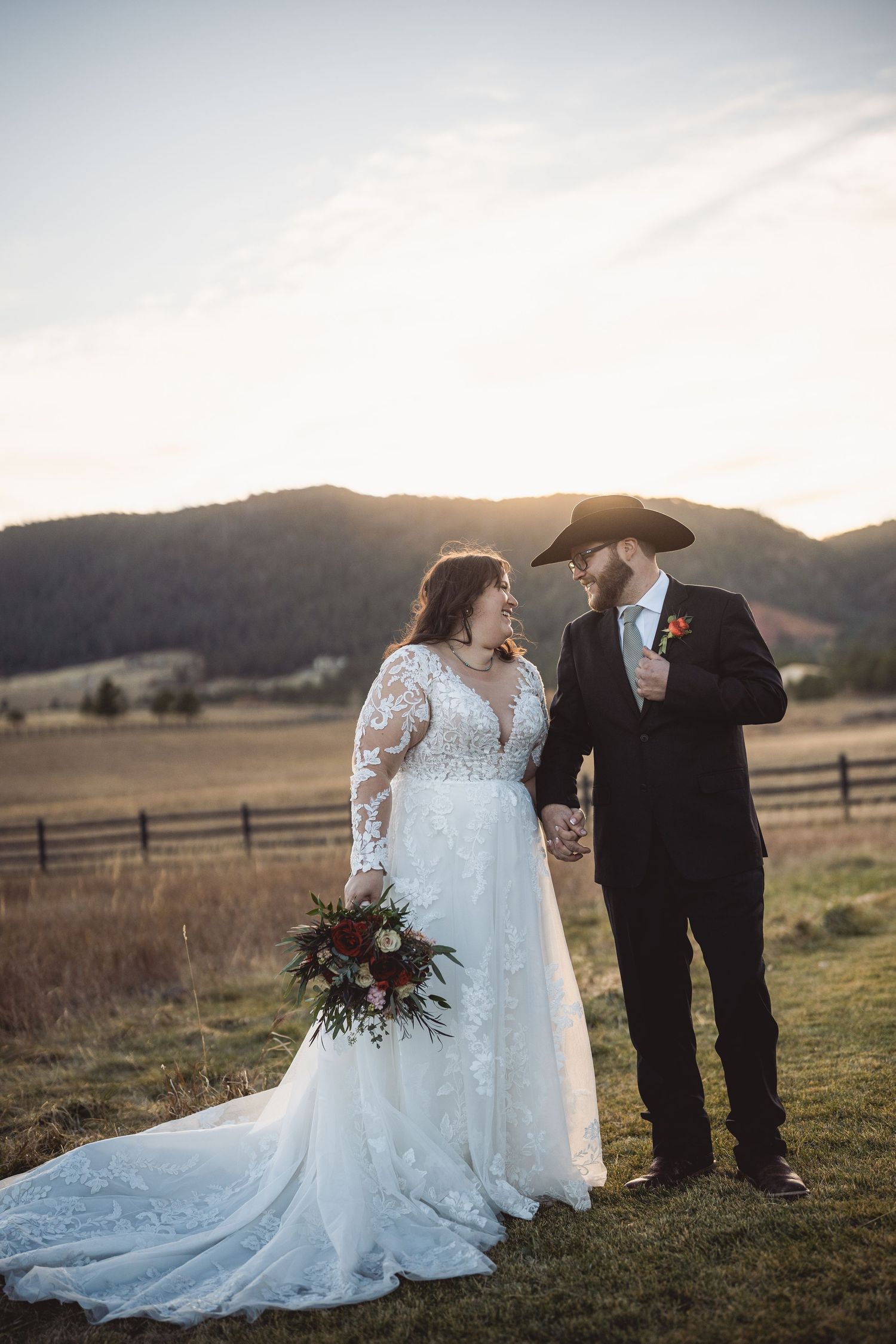 Elegant wedding photography session in a field with mountain views and wooden fence at sunset.