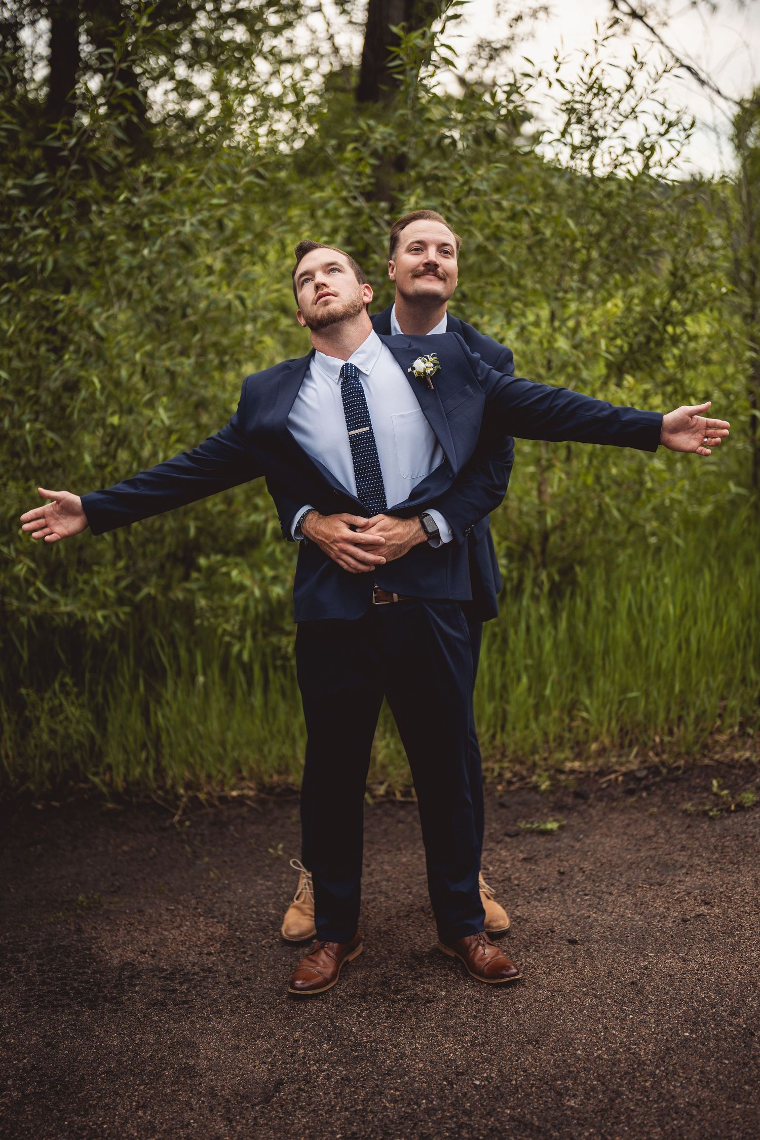 Two people in suits performing dramatic poses on dirt path with greenery background.