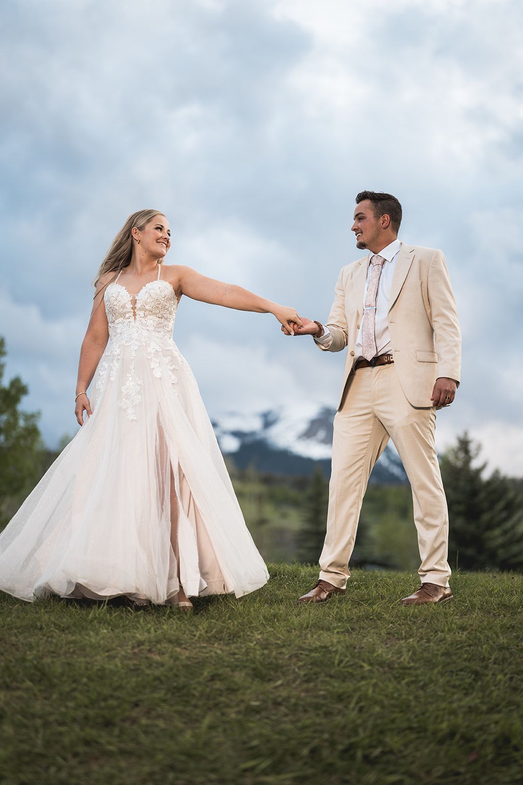 Bride and groom holding hands outdoors on grass with mountain backdrop, bride in lace gown, groom in tan suit.