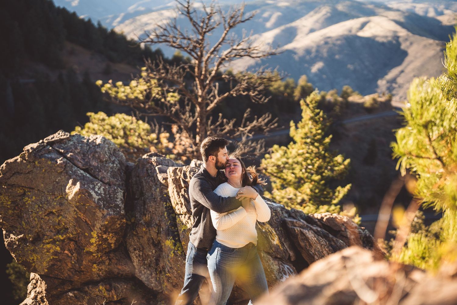 Couple embracing romantically on rocky mountain overlook surrounded by pine trees and scenic valley views.