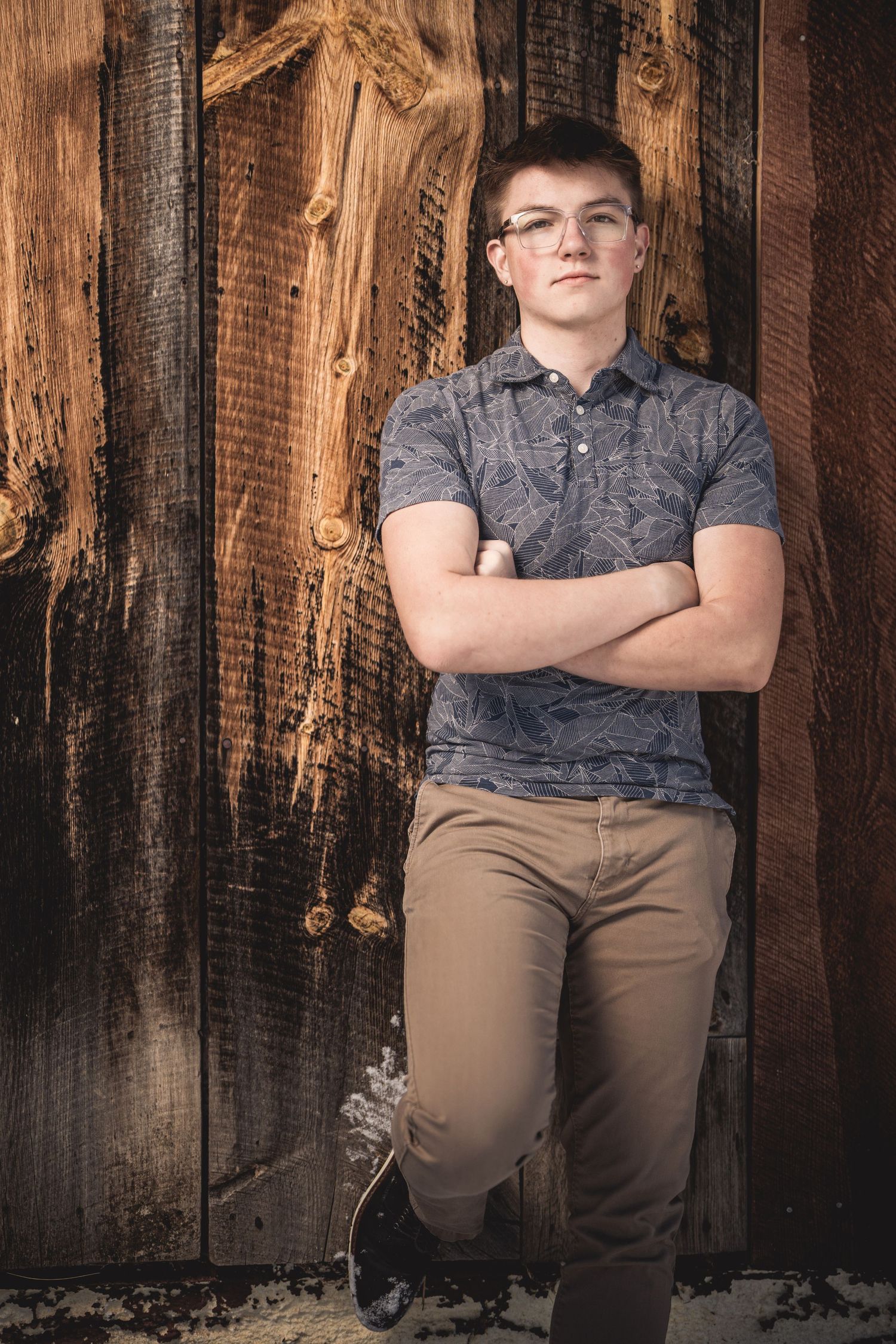 Teen boy with glasses leaning against rustic wood wall, arms crossed, wearing gray polo and khaki pants.