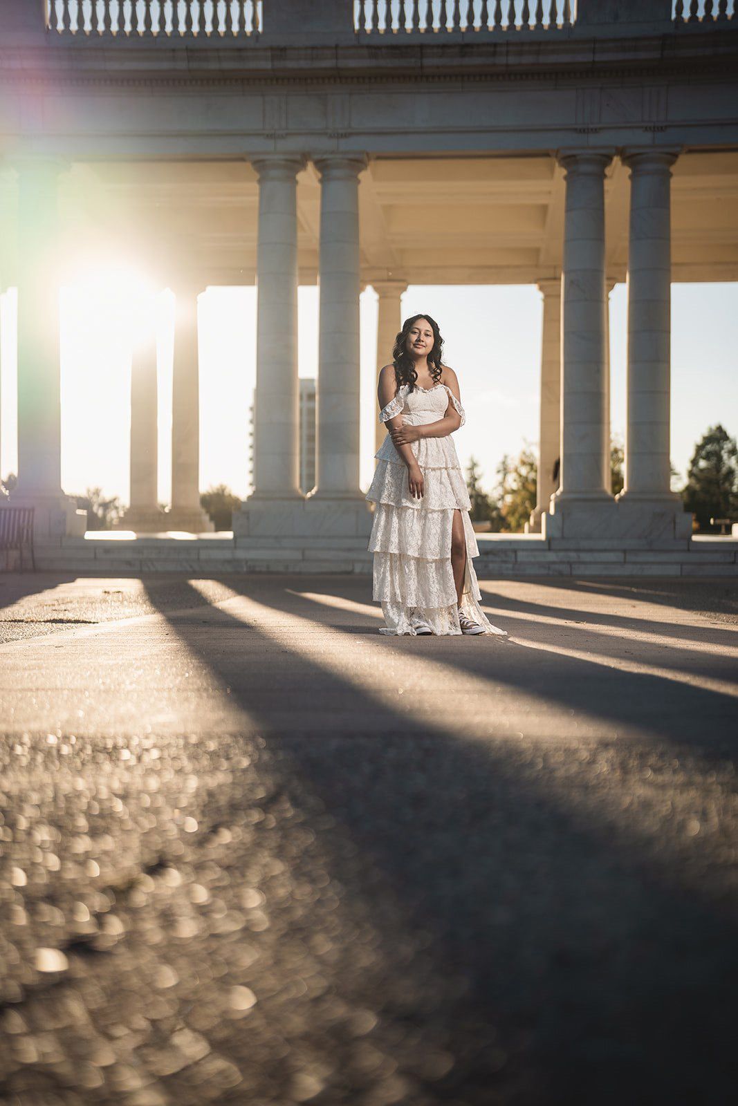 Woman in white dress poses before sunlit classical columns at golden hour.