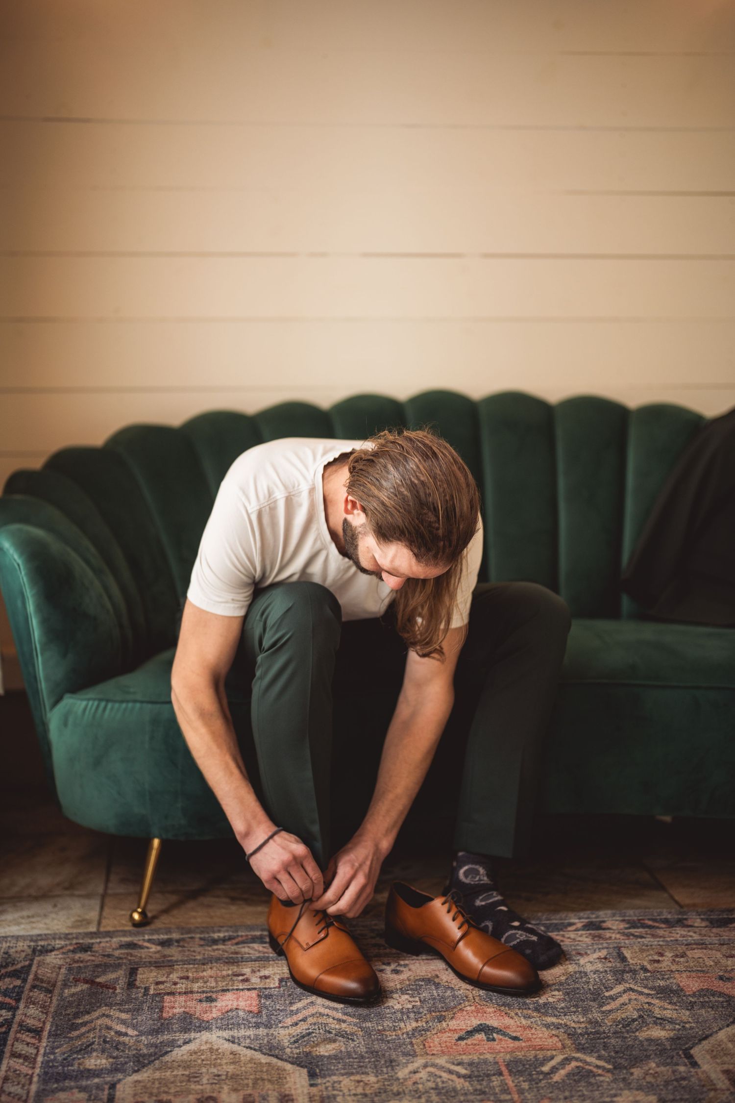 Person sitting on green velvet couch tying brown leather dress shoes.