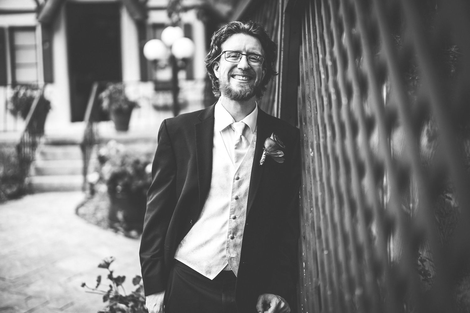 A groom in a black suit and light vest smiles while leaning against a textured wall.