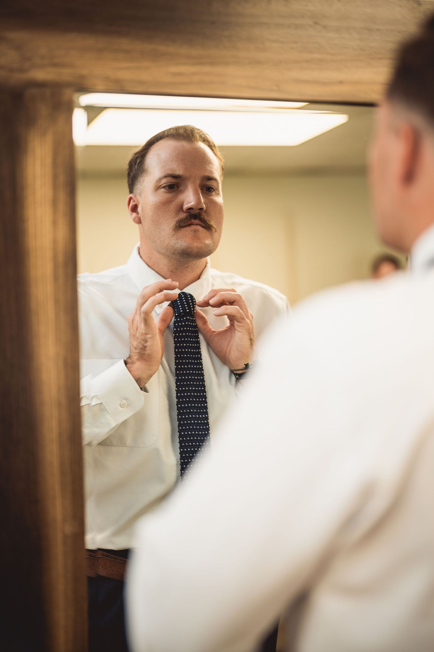 A man adjusts his tie while looking in a mirror surrounded by wooden frames.