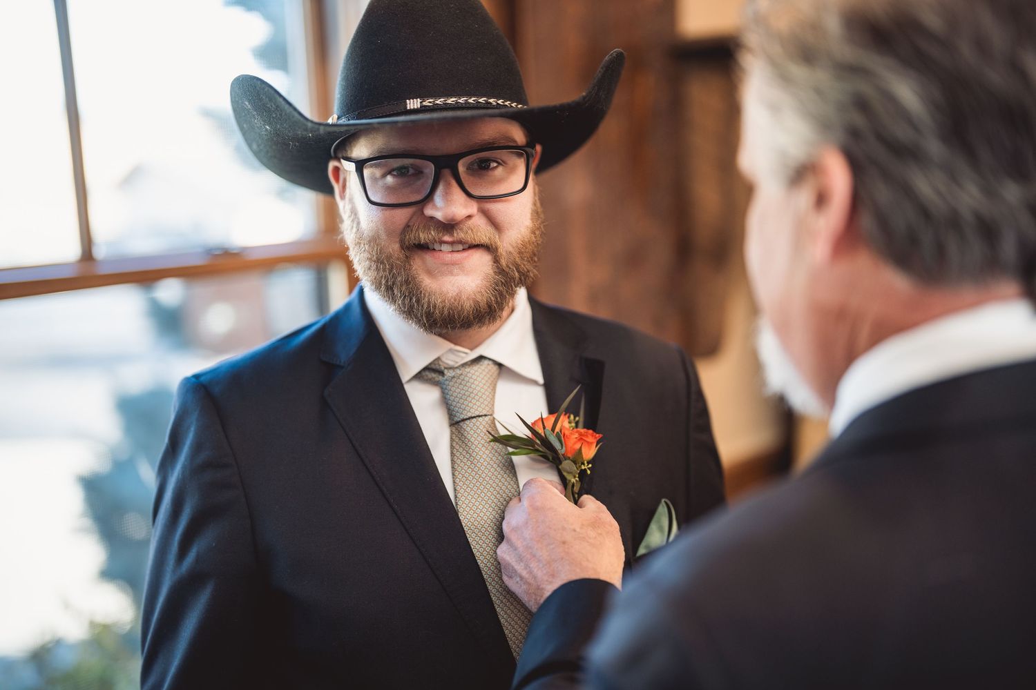 Groom in cowboy hat and glasses adjusts his tie while standing in warm indoor light.