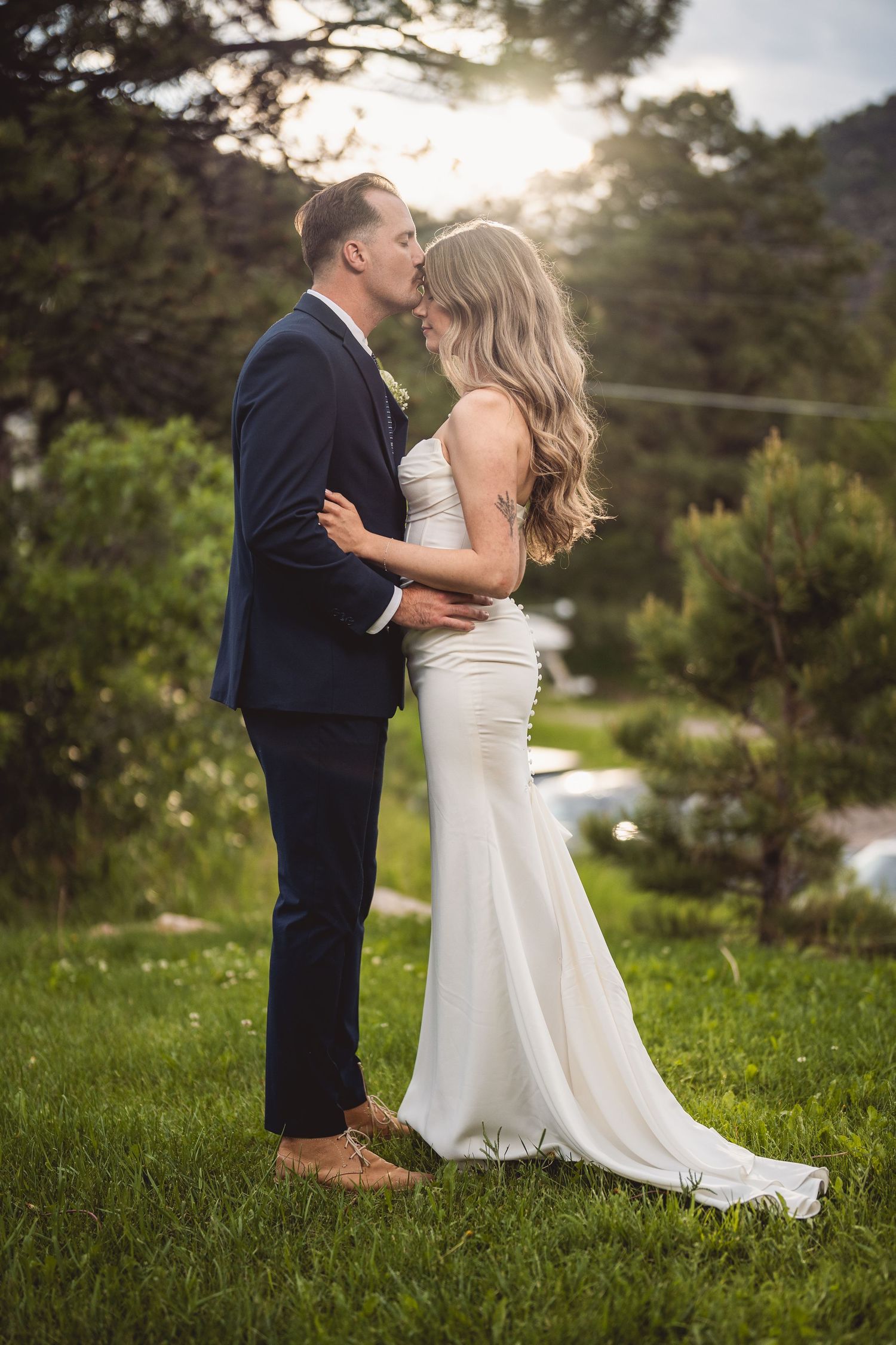A romantic wedding couple embraces outdoors with sunlight streaming through pine trees in the background.