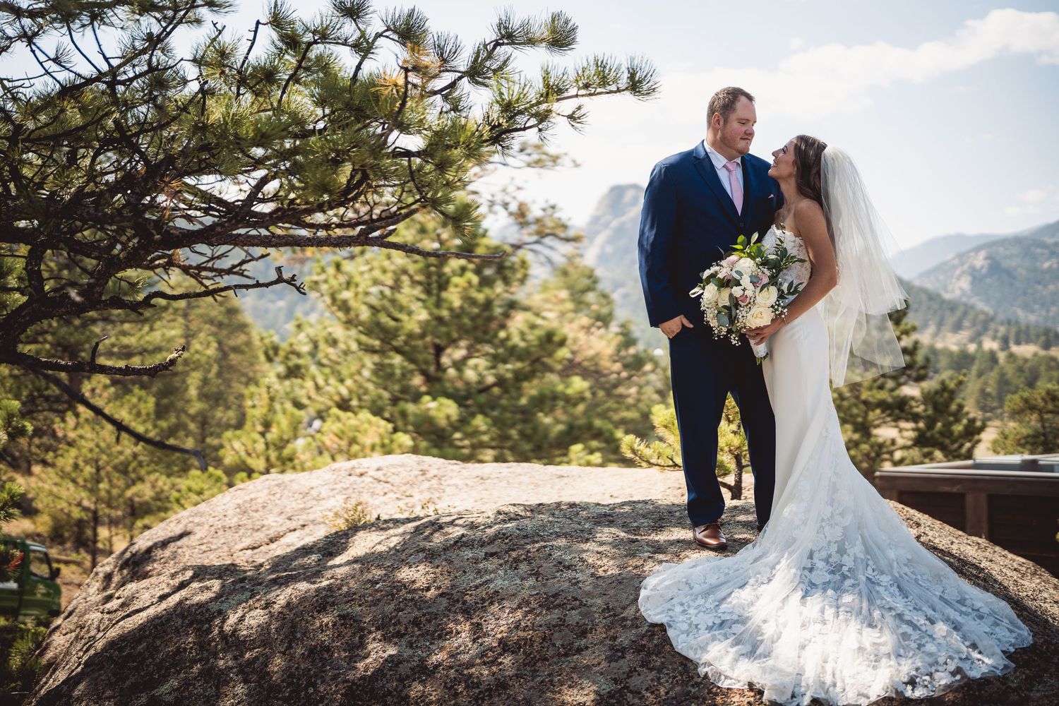 Bride and groom stand on rocky mountain overlook surrounded by pine trees, bride holding white bouquet in lace gown.