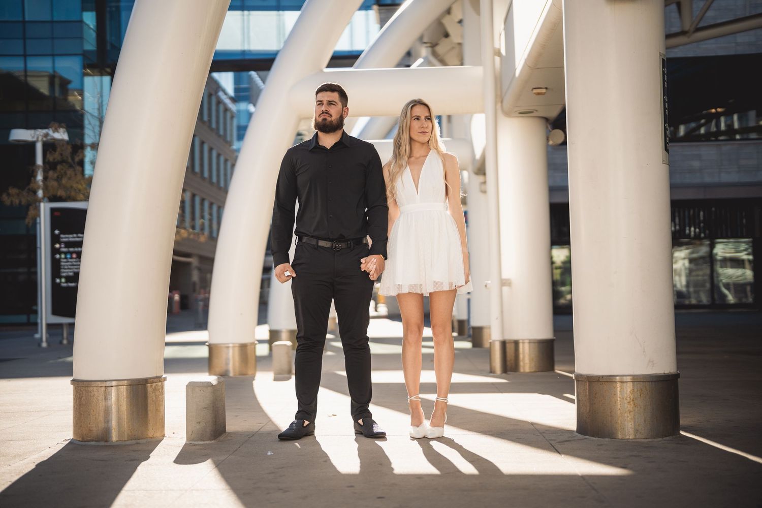 A couple poses outdoors among white architectural columns, man in black outfit, woman in white mini dress.
