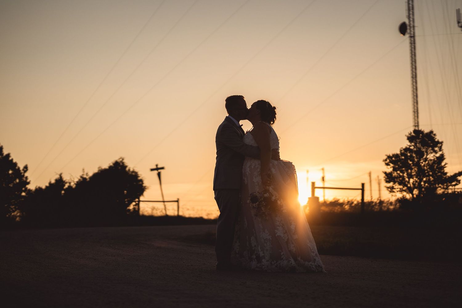 Silhouette of a bride and groom kissing at sunset with warm golden sky backdrop at an outdoor wedding.