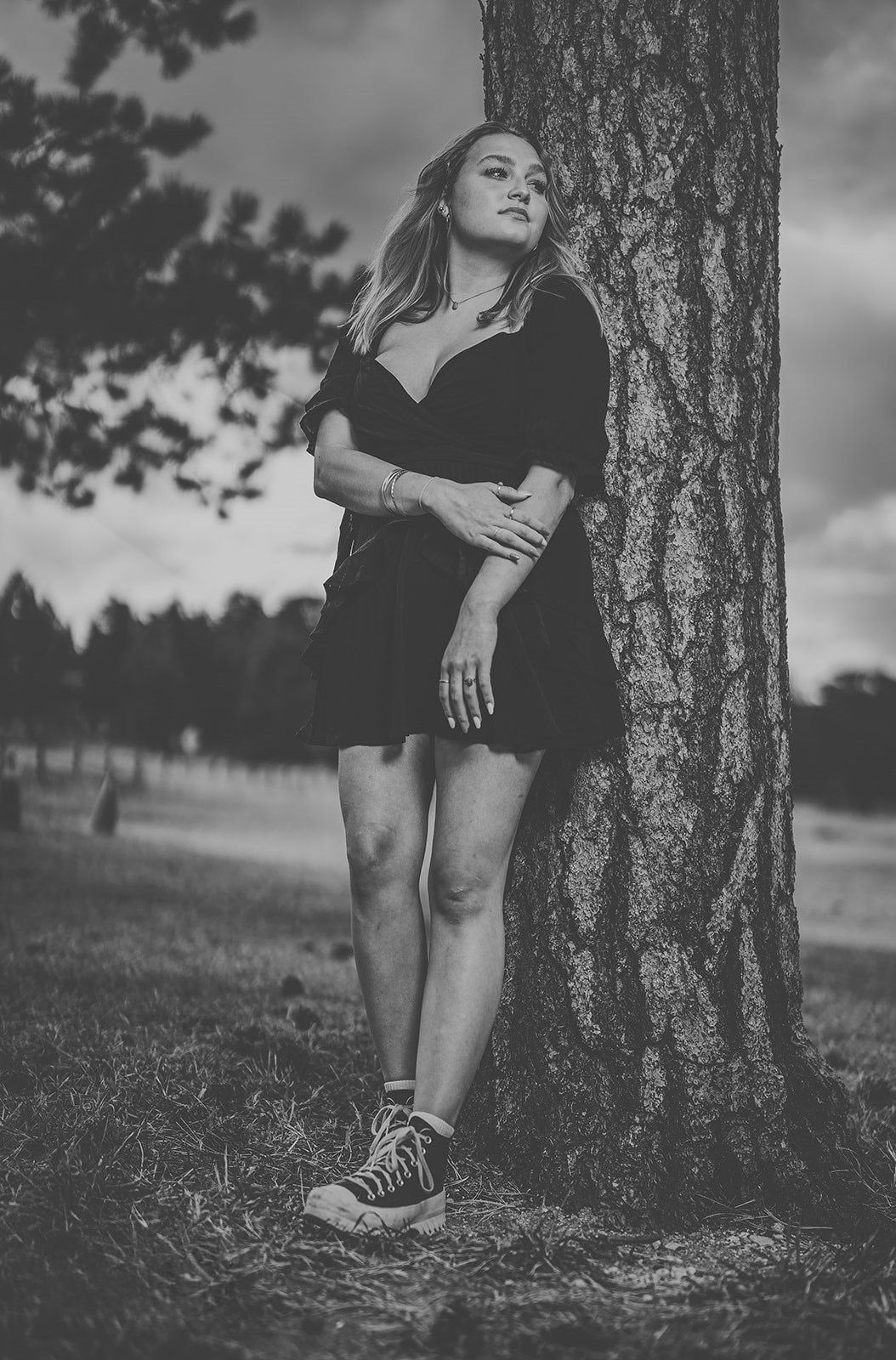 Black and white portrait of blonde woman leaning against tree in short black dress.
