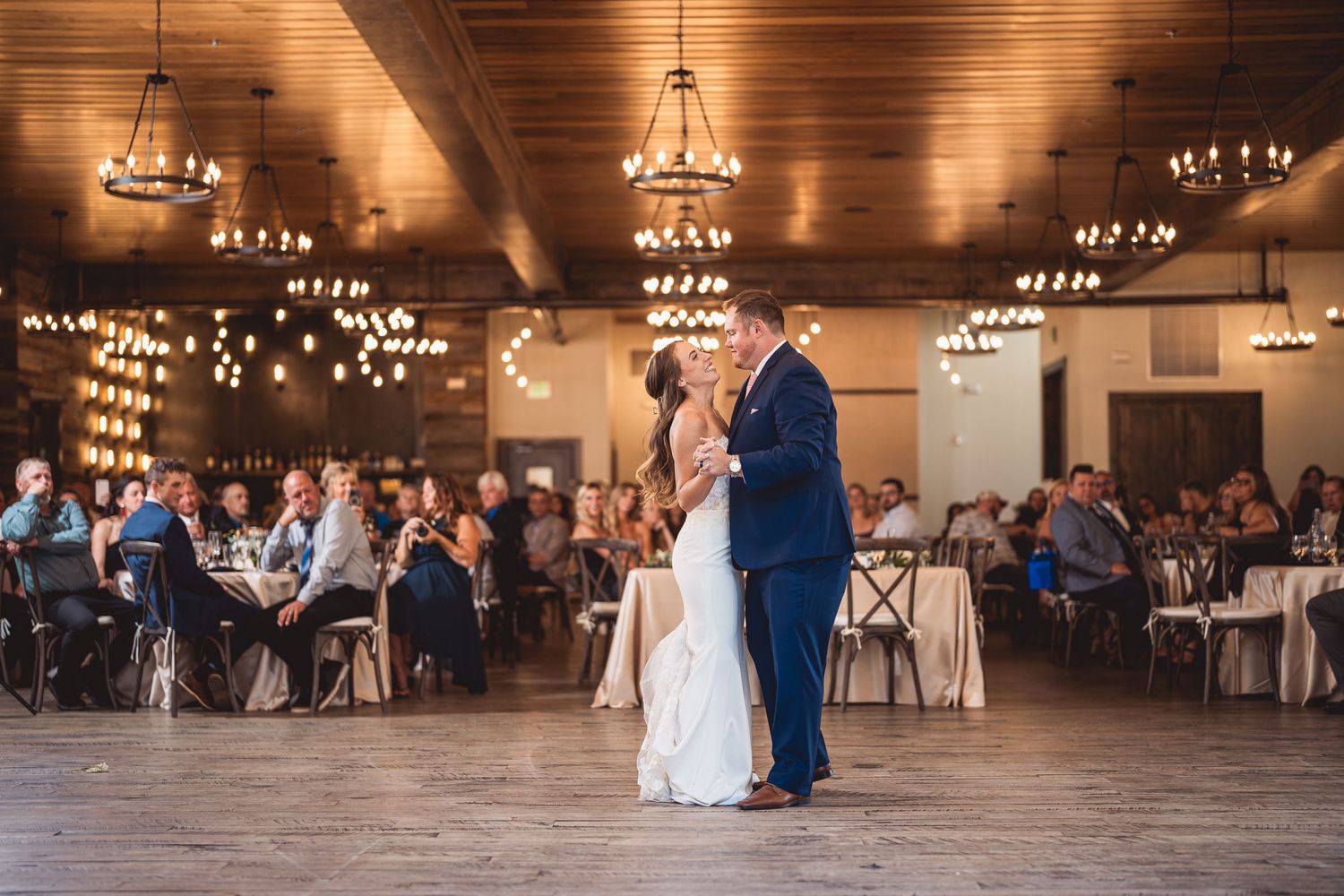 Bride and groom share first dance in elegant rustic wedding venue with chandeliers and guests seated at tables.