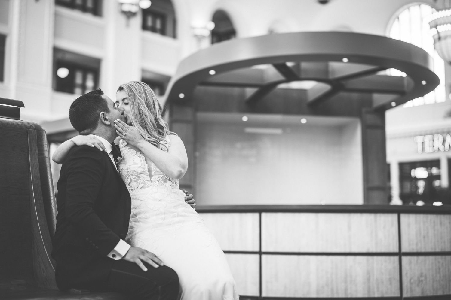 Black and white wedding photo of bride and groom sharing a romantic kiss while seated in an elegant venue.