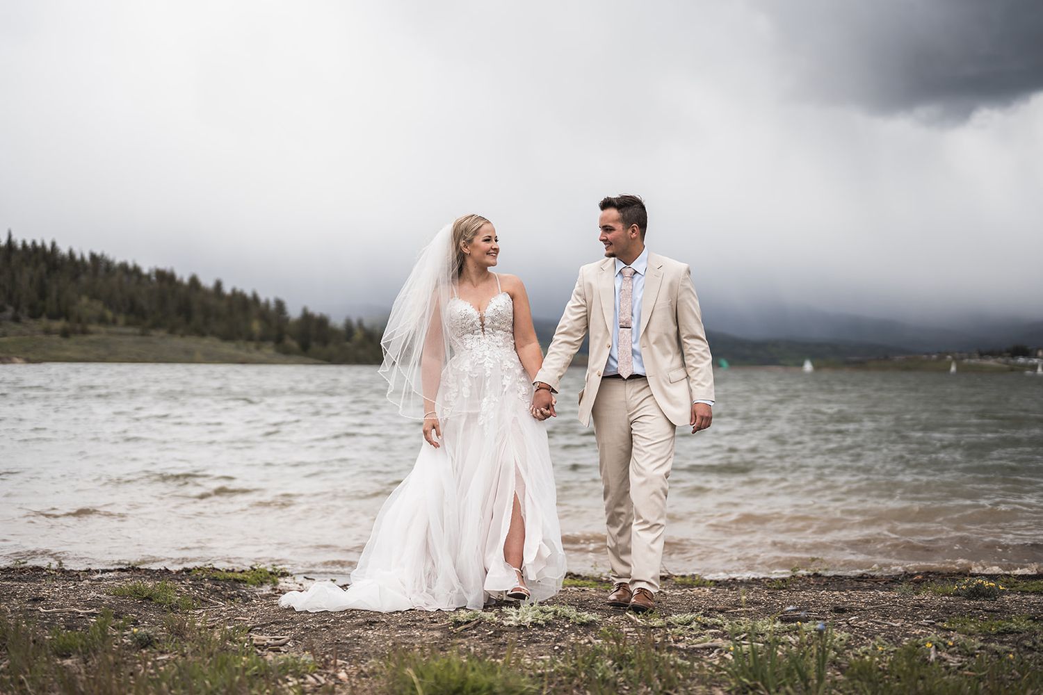 Bride and groom holding hands walking along a lakeside shore with stormy mountain skies in the background.