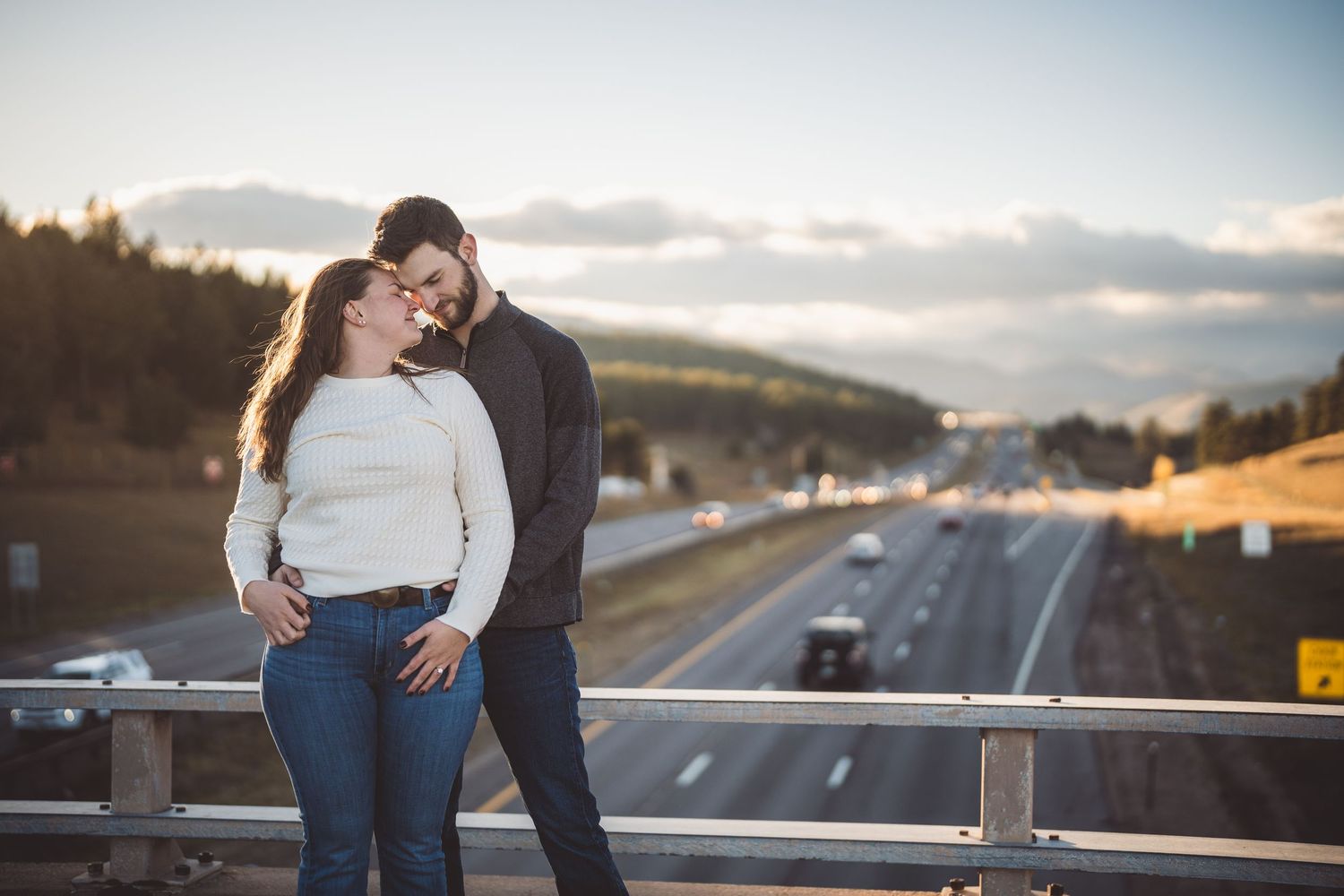 Couple sharing a romantic moment on a bridge overlooking a busy highway at golden hour.