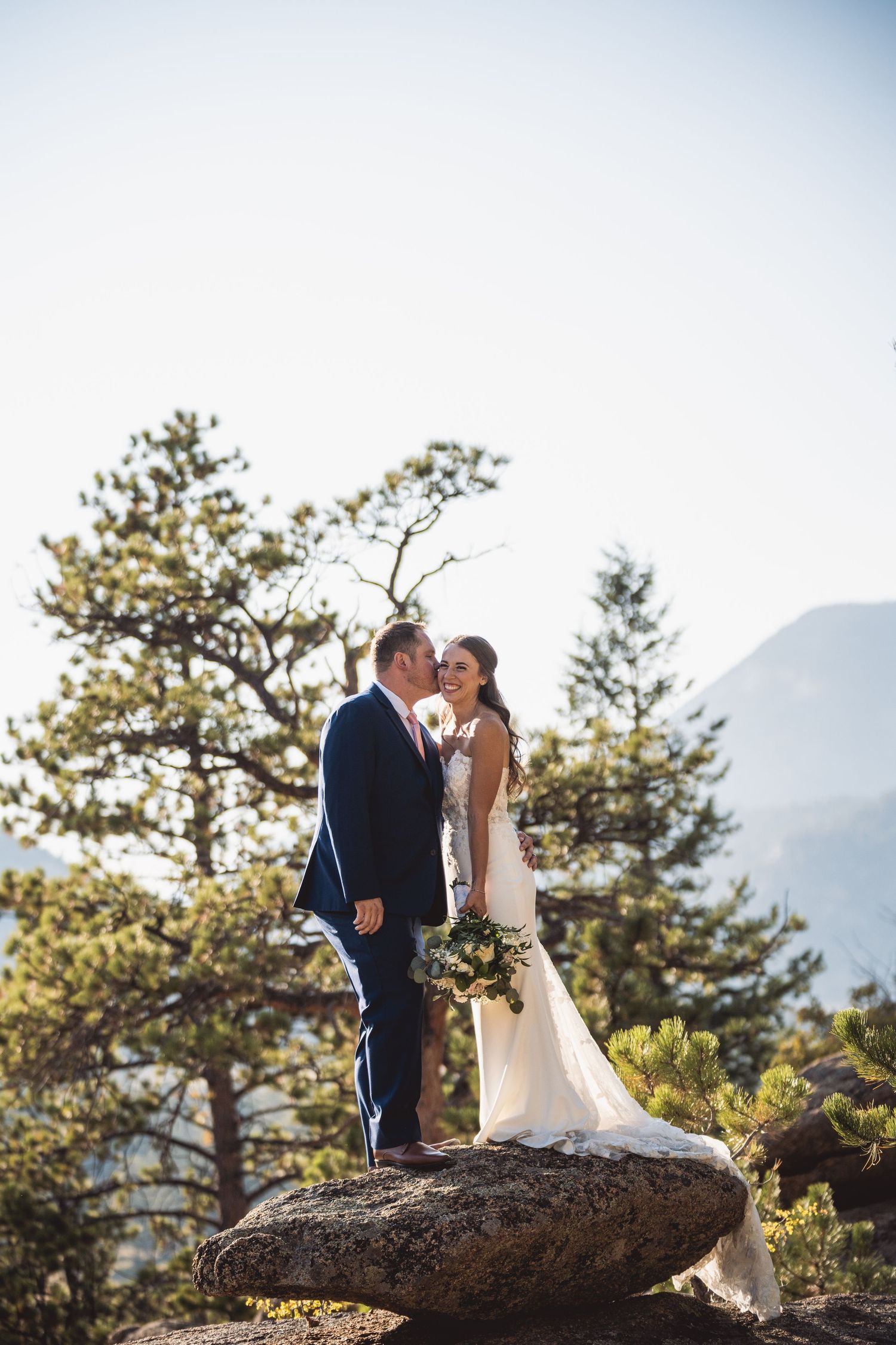 Bride and groom standing on a large rock with pine trees and mountains in the background on their wedding day.
