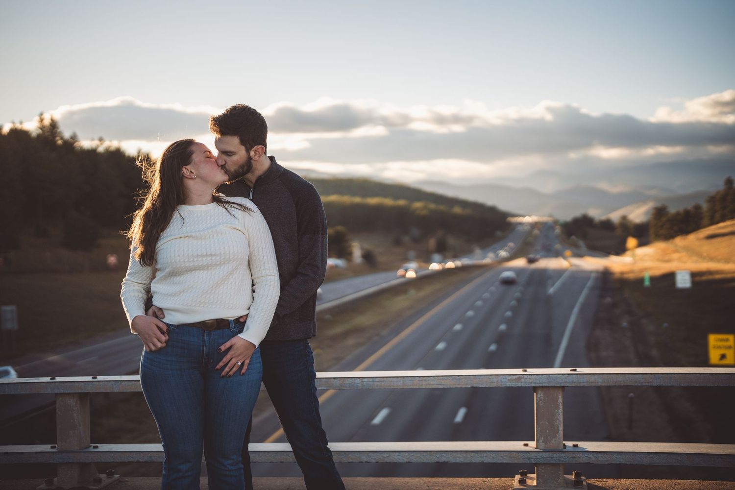 Couple sharing a romantic kiss on a bridge overlooking a highway with mountain views at sunset.