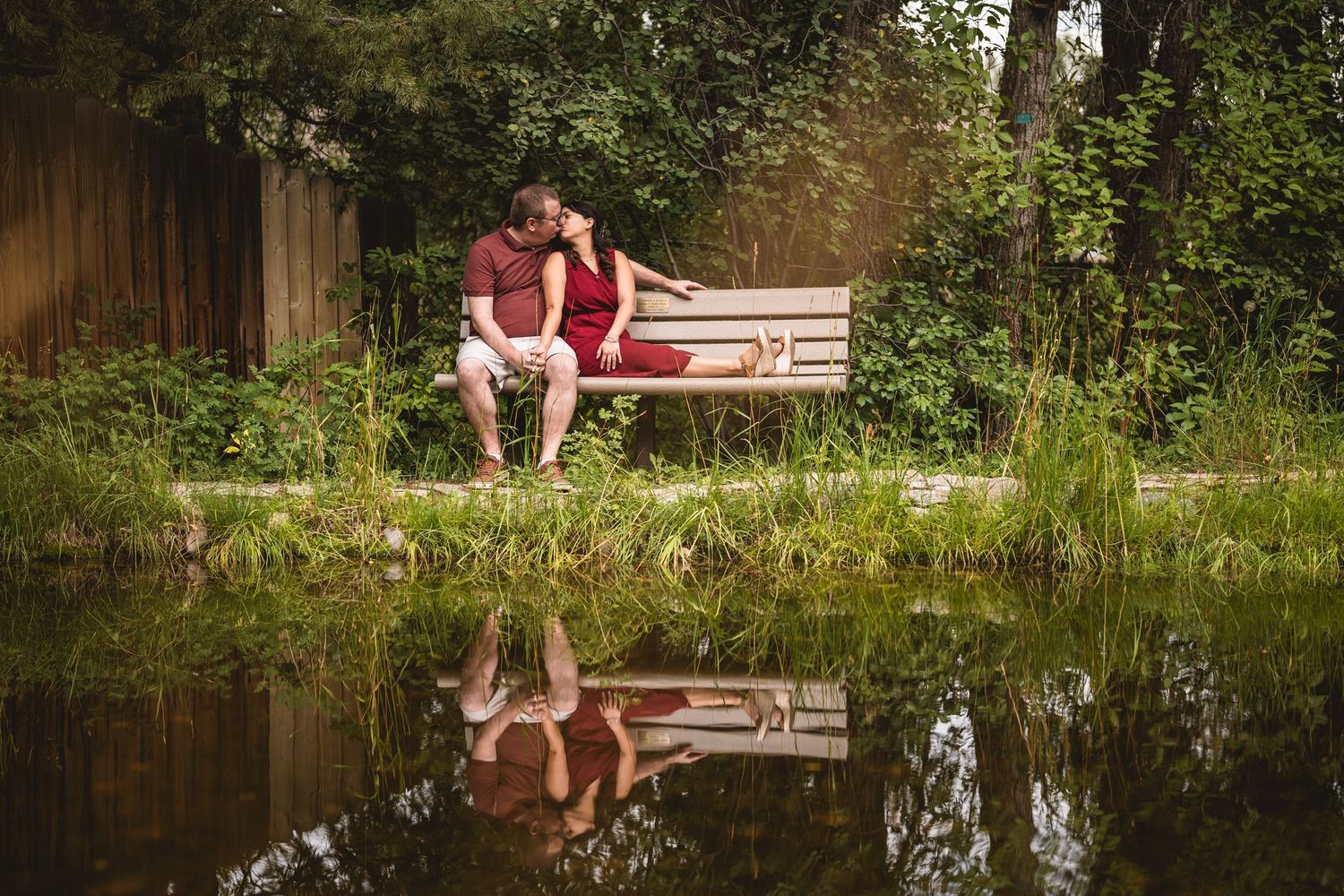 A romantic couple sits together at the edge of a serene pond surrounded by lush greenery.