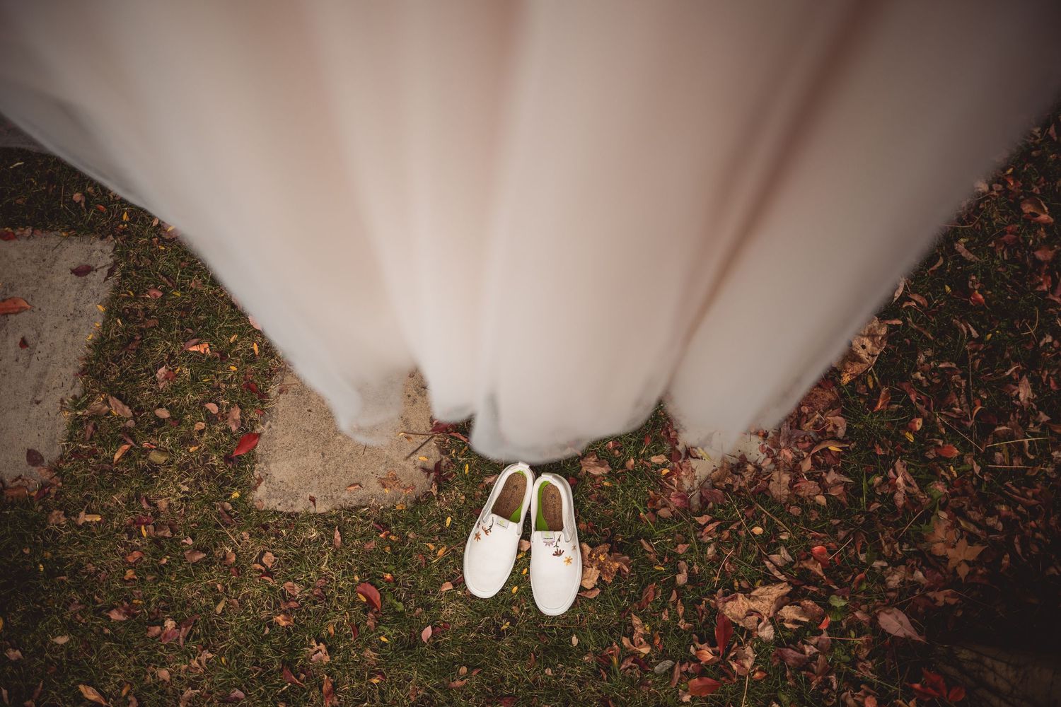 White bridal shoes and dress hem photographed against fallen autumn leaves.