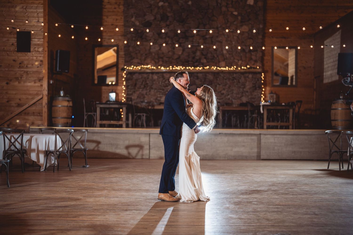 A romantic first dance on a rustic wooden floor under twinkling string lights in a barn venue.
