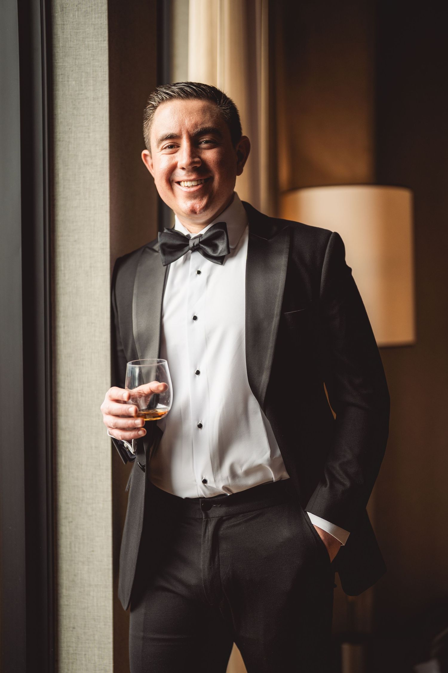 Person in formal black tuxedo holding wine glass by window with warm lighting.