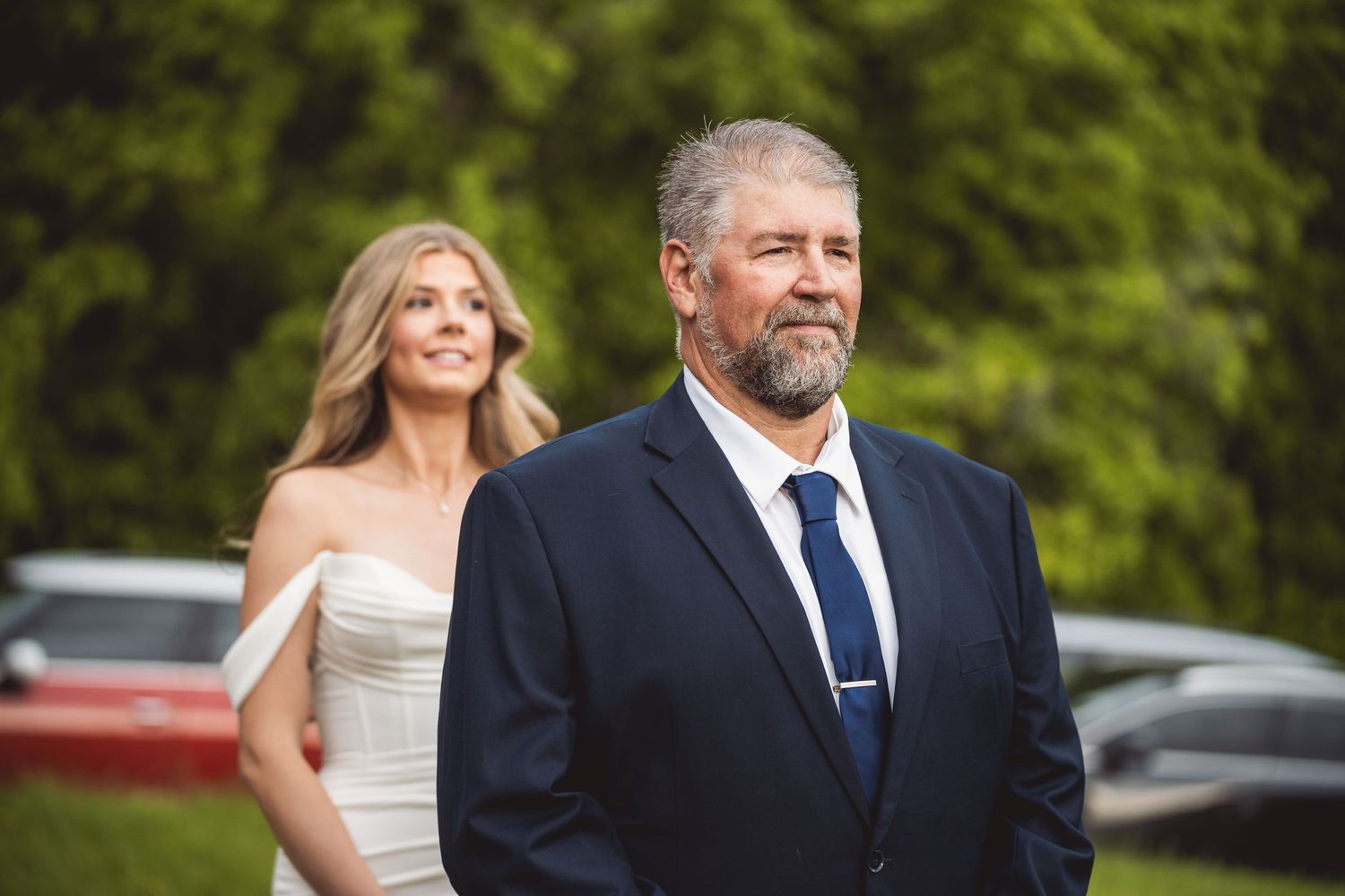 A couple stands close together outdoors with lush greenery in the background.