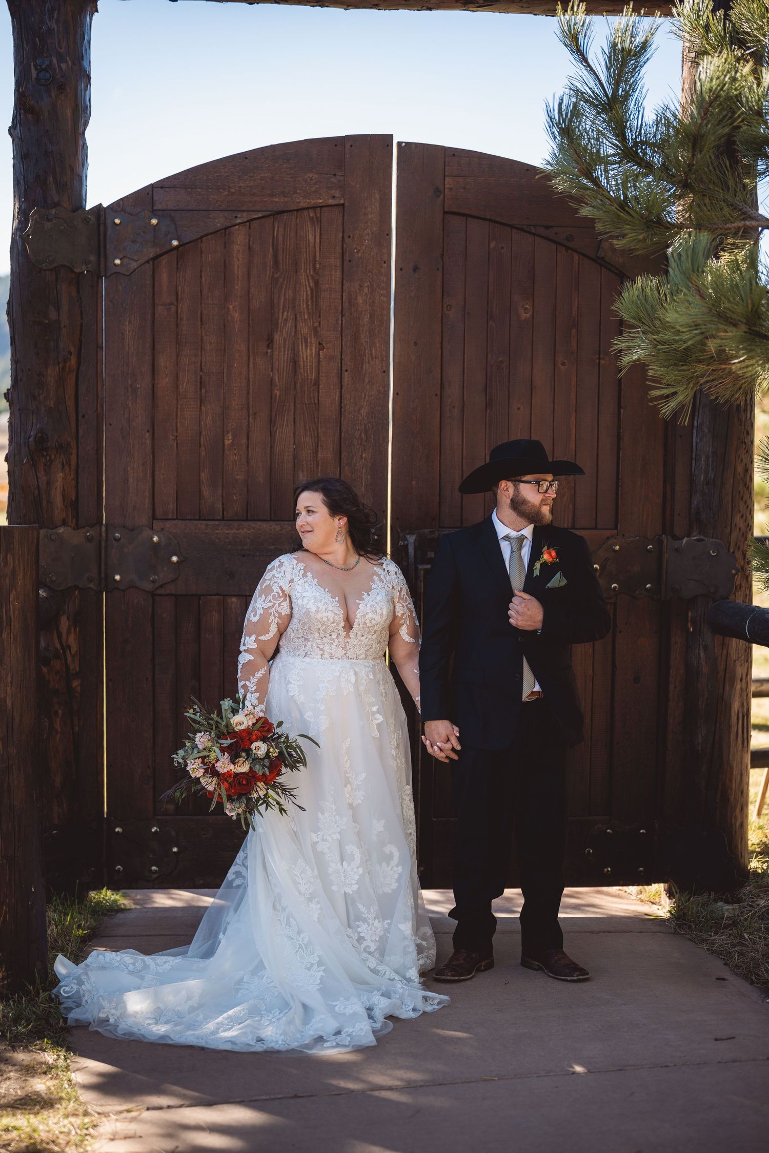 A bride in lace wedding dress stands with others by rustic wooden gates outdoors.