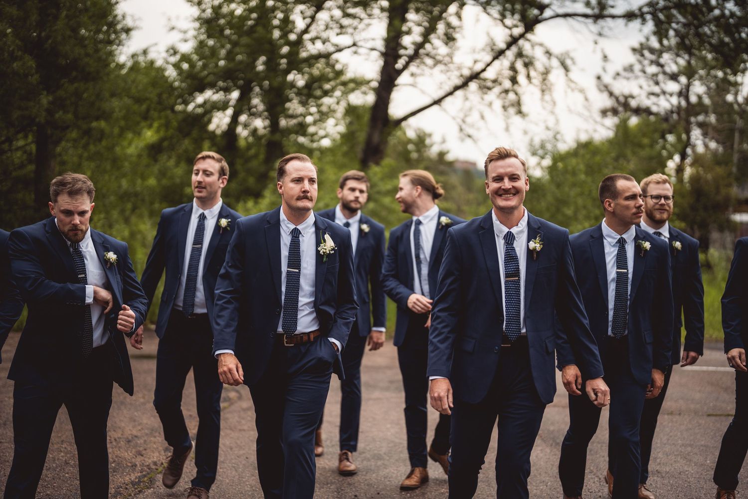 Wedding party in navy suits walking and laughing together on a garden path.
