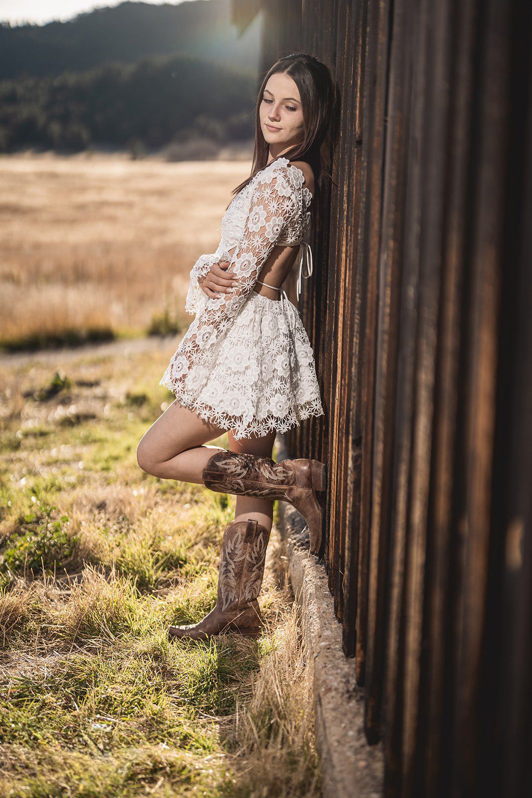 Young woman in white lace dress and cowboy boots leaning against rustic wooden wall.