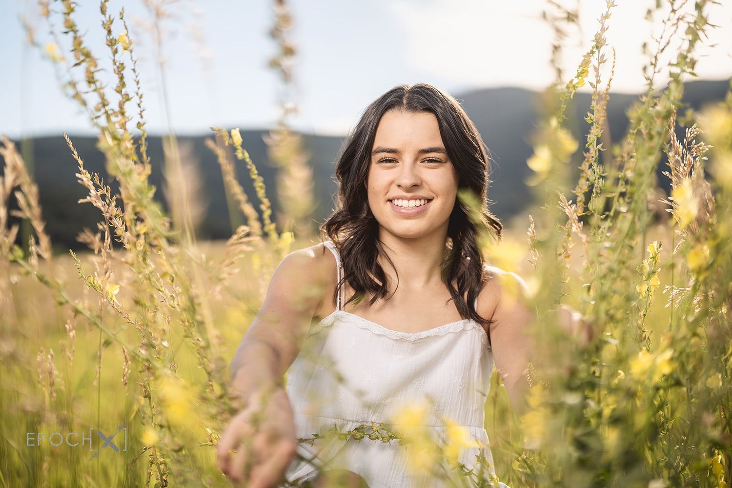 Smiling young woman in white dress standing among tall wildflowers in a sunny field with mountains in the background.