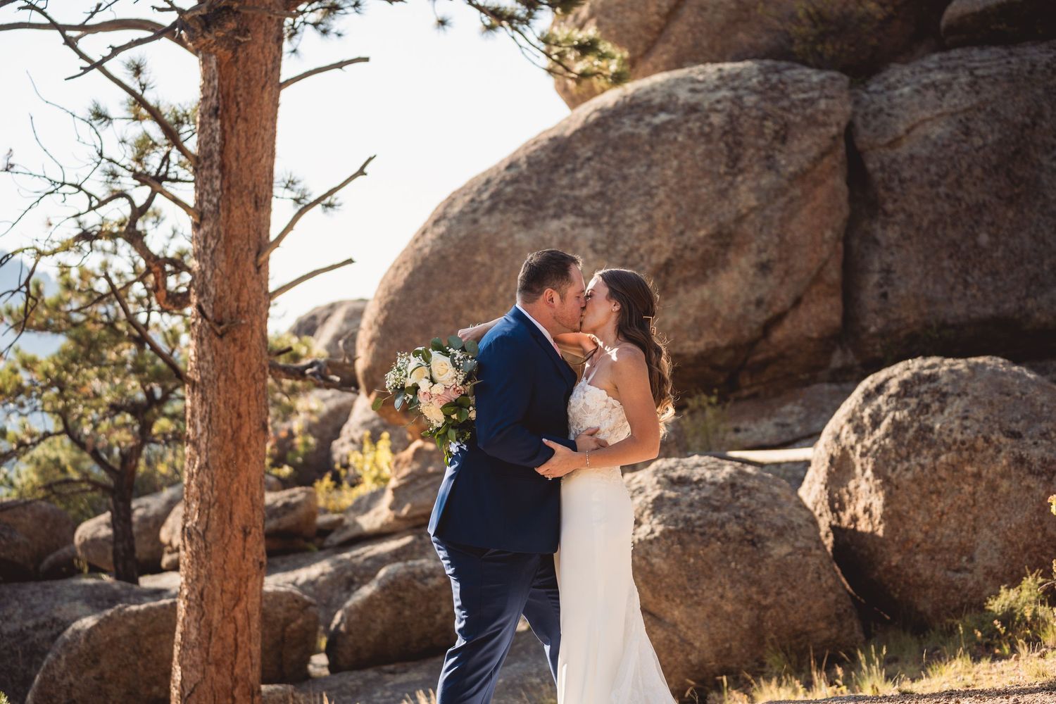 A couple shares a romantic kiss surrounded by large boulders and pine trees during their outdoor wedding ceremony.