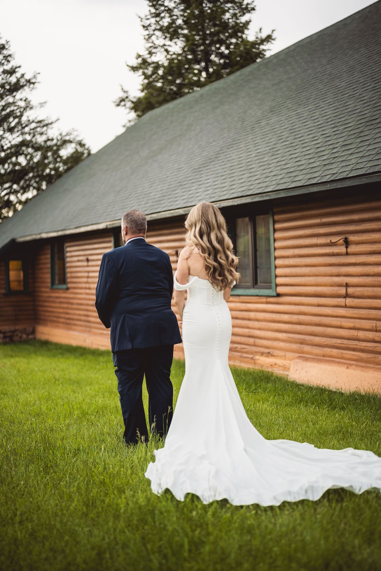 Bride and groom walking by log cabin building with flowing white wedding dress trailing on green grass.