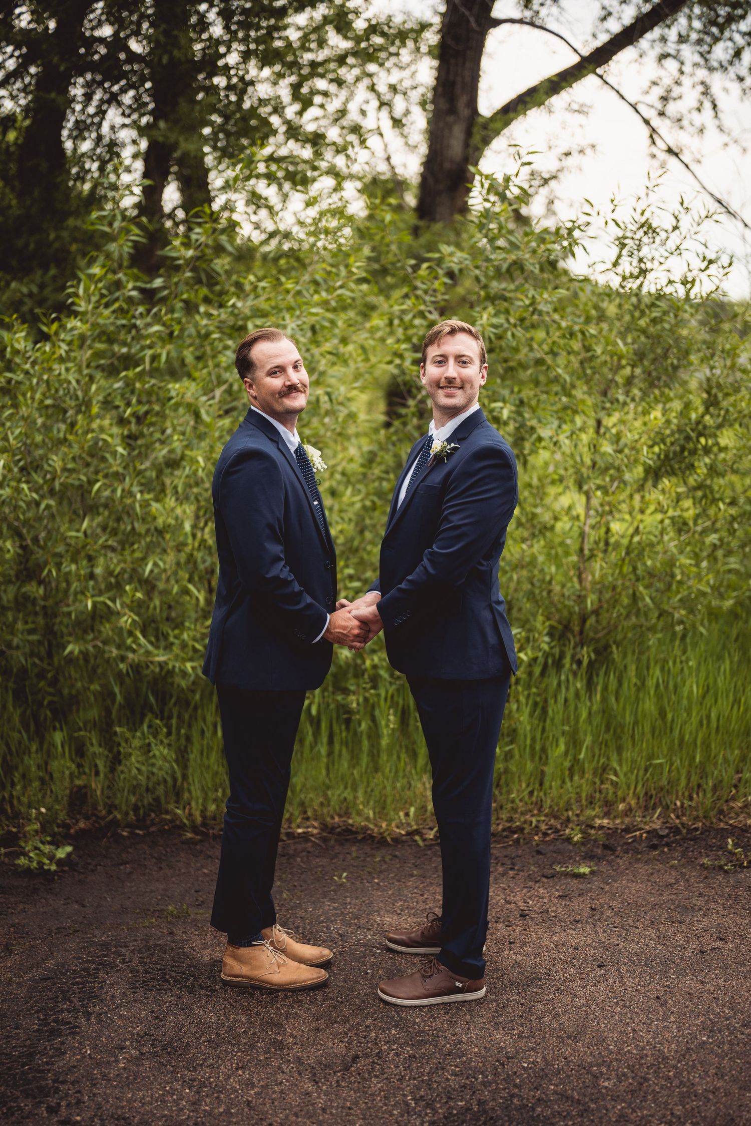 Two groomsmen in navy suits sharing lighthearted moment on garden path.