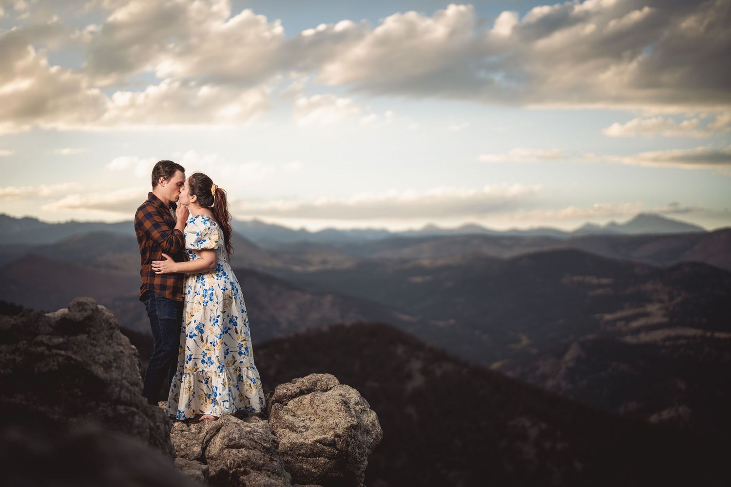 Couple sharing a romantic kiss on a rocky mountain summit with stunning panoramic views and dramatic cloudy sky.