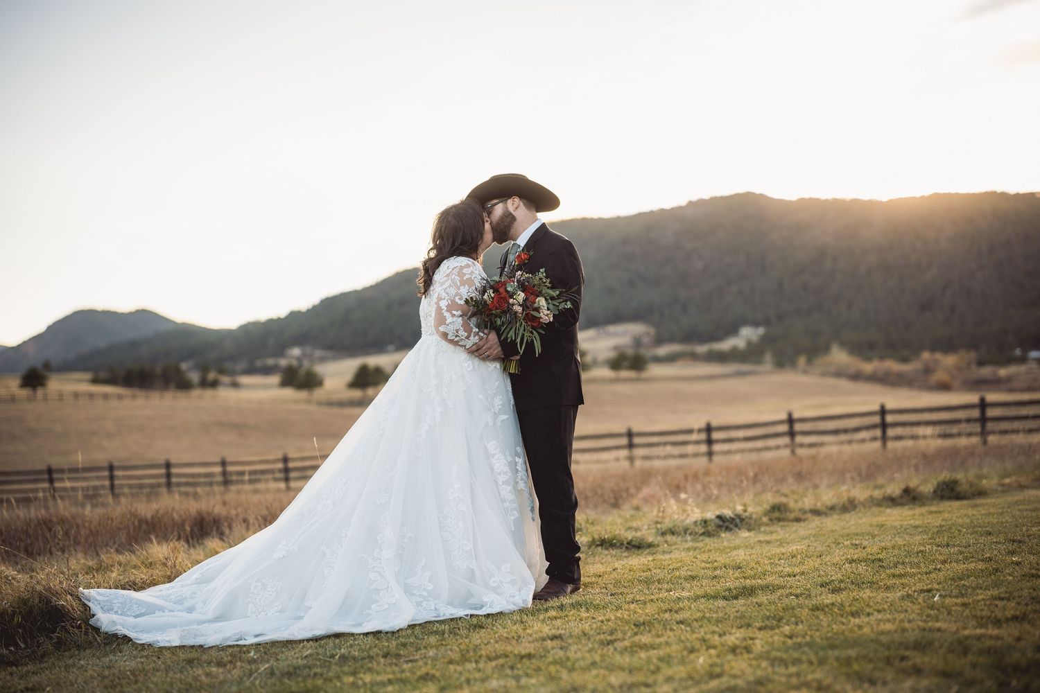 Bride and groom share a romantic kiss at sunset in a rustic mountain ranch setting, with rolling hills and wooden fences.